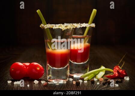 Dos vasos de tiro con cóctel Bloody Mary, apio, especias y tomates maduros sobre fondo de madera oscura Foto de stock