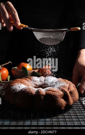La mano de la mujer rocía azúcar glaseado sobre la torta fresca del bundt. Fondo oscuro. Foto de stock