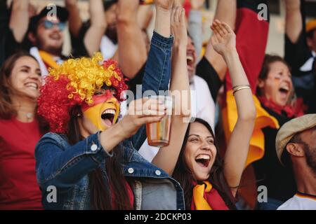 Mujer en la zona de fans con una peluca y cara pintada en colores de bandera alemana sosteniendo un vaso de cerveza y animando con multitud de espectadores en el evento deportivo. Alemania