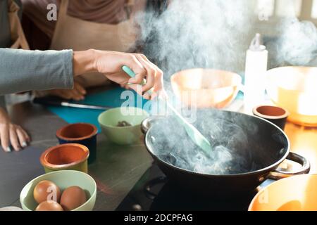Mano de mujer joven con espátula resistente al calor mezclando ingredientes en sartén Foto de stock