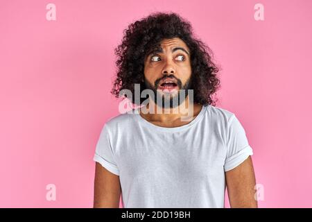 joven árabe está sorprendido, mostrar sorpresa y expresión asombrada. guapo chico con el pelo rizado largo y barba permanecer en shock, aislado sobre fondo rosa Foto de stock