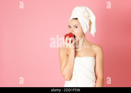 se mofa, retrato de una joven con una toalla en la cabeza y en su cuerpo mordiendo una granada roja aislada sobre un fondo rosa Foto de stock