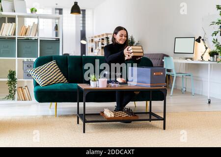 Sonriente hermosa mujer recogiendo libros en la caja en la mesa de café en apartamento loft