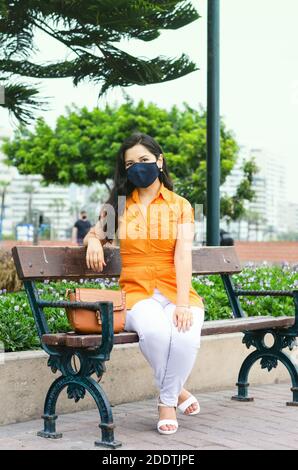 Mujer que usa una máscara para protegerse del covid19 sentada en un parque. Mujer que se relaja en un parque durante la primavera usando una máscara protectora, sentada en un banco bajo el sol durante la pandemia Foto de stock