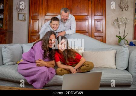 Feliz familia teniendo una videollamada con amigos, saludando la pantalla del portátil - Padre, madre, hija e hijo en el sofá en casa haciendo un video chat w