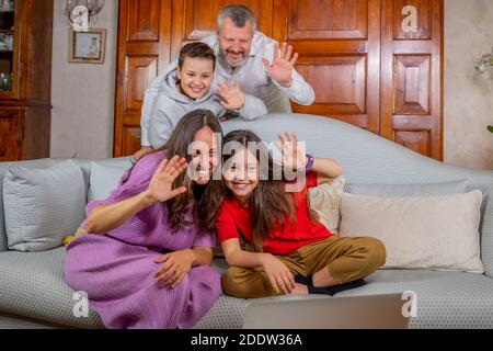Feliz familia tener una videollamada con amigos, saludando la pantalla del portátil - Padre, madre, hija e hijo en el sofá en casa haciendo una charla de vídeo