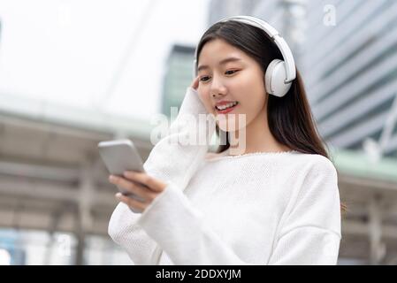 Feliz y sonriente chica asiática con auriculares y mirando el smartphone mientras escucha música en streaming contra el fondo de la construcción de la ciudad Foto de stock