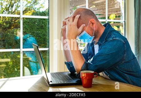 Estresado hombre sentado junto a su ordenador portátil con las manos en la cara mientras llevaba una máscara. . Foto de alta calidad Foto de stock