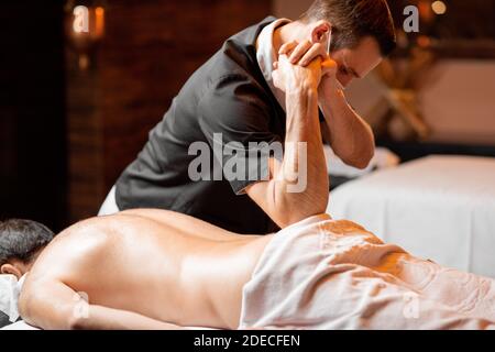 Masajista profesional en máscara facial haciendo un masaje profundo a un cliente masculino en el salón Spa. Negocio durante el concepto epidémico Foto de stock