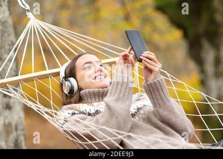 Mujer feliz con auriculares y teléfono inteligente escuchando música tumbado en una hamaca en las vacaciones de otoño Foto de stock