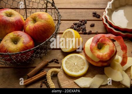 Elaboración de un pastel de manzana: Mesa de madera con una cesta de manzanas rojas, una pelada y algunas rebanadas de manzanas, limón, canela y pasas. Foto de stock