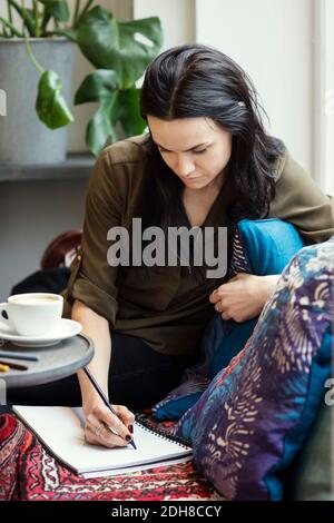 Mujer blogger concentrada escribiendo en espiral portátil mientras se sienta sofá