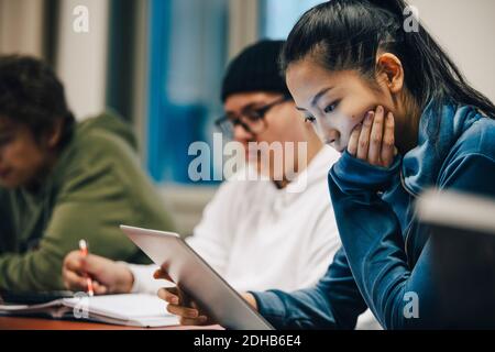 Niña adolescente con la mano en la barbilla usando tableta digital mientras sentado por compañeros de clase en la escuela secundaria