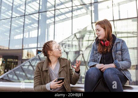 Niña adolescente con teléfono móvil hablando con un amigo mientras está sentada contra la construcción