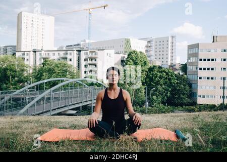 Mujer joven meditando en el campo contra los edificios de la ciudad
