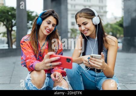 Alegre hipster estudiantes en trajes de moda surfing redes sociales en teléfonos móviles y escuchar música con auriculares mientras sentado toget Foto de stock