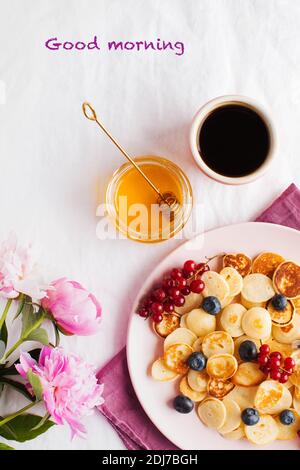 Pequeñas panqueques con bayas, miel, flores, café y las palabras Buenos días. Concepto de desayuno Fotografía de stock