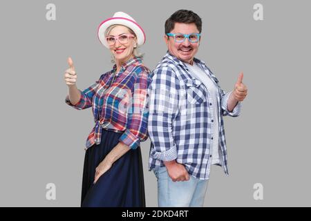 Par de amigos, un hombre adulto y una mujer en casual camisa a cuadros juntos la espalda, mostrando Thumbs up, gran sonrisa y mirando a la cámara Foto de stock