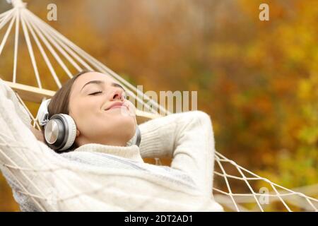 Mujer relajada escuchando música con auriculares tumbados en la hamaca en otoño en un bosque Foto de stock