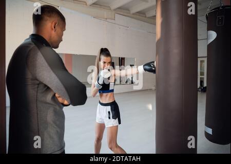 Un entrenador estricto observa a su estudiante de kickboxer practicar un ponle en una bolsa de perforación en una amplia sala de entrenamiento Foto de stock