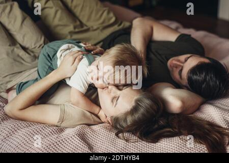Feliz familia con un pequeño hijo acostado y jugando en la cama en casa