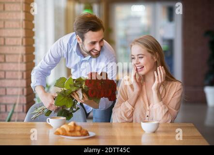 Hombre milenial cariñoso sorprendiendo a su novia, dándole un ramo de flores en el café Foto de stock