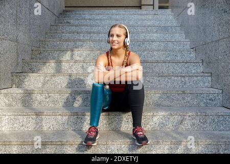 Sportswoman sonriente con pierna protésica con auriculares sentados con los brazos atravesado por la escalera Foto de stock