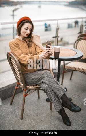 Encantadora mujer joven francesa usando su smartphone tomando selfie mientras una taza de café. Mujer francesa con boina roja sentada en la terraza de Fotografía de - Alamy