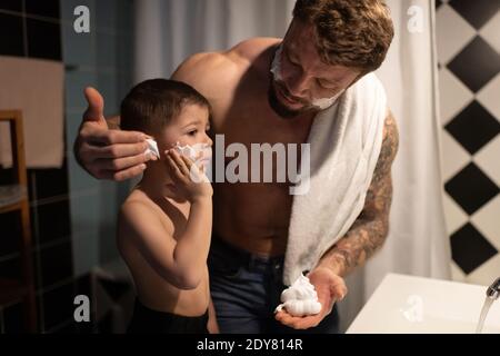 Un hombre con barba adulto que ayuda a un niño pequeño a impregnar espuma mejillas mientras se prepara para afeitarse la cara por la mañana juntos Foto de stock