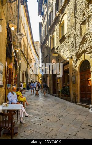 Vislumbre de un estrecho callejón en el casco antiguo con gente y turistas caminando y sentados en un café al aire libre en verano, Volterra, Pisa, Toscana, Italia