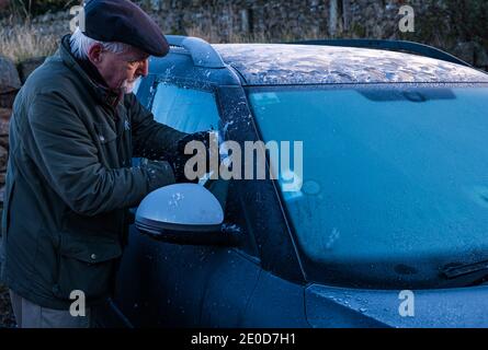 Hombre mayor raspar hielo del parabrisas del coche en invierno con patrones de escarcha en el techo del coche, Escocia, Reino Unido Foto de stock
