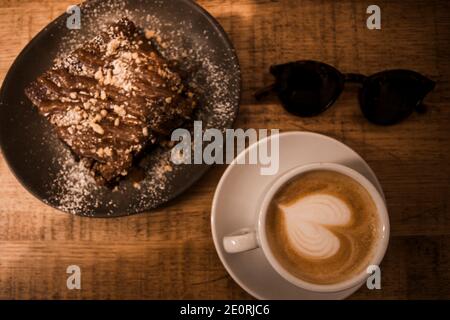 Plato con una rebanada de deliciosa tarta de chocolate casera sobre una mesa de madera. Pastel de brownie casero con un sabroso capuchino italiano fresco para un desayuno saludable Foto de stock