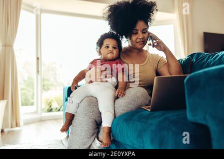 Madre africana sentada con un bebé en el sofá y hablando por teléfono y trabajando con un ordenador portátil.
