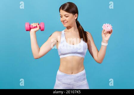 Mujer atlética positiva con cuerpo delgado que sostiene en manos de color rosa mancuerno y donut con esmalte rosa, haciendo la elección en favor del deporte y el estilo de vida saludable Foto de stock