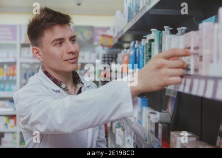 Alegre y guapo farmacéutico masculino haciendo inventario, organizando productos en las estanterías de la farmacia. Encantador químico joven que trabaja en la farmacia. Foto de stock