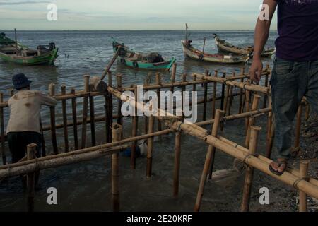 Hombres construyendo la estructura costera utilizando postes de bambú, un edificio de pilotes sobre el agua del mar. Los barcos de pesca se pueden ver en el bakground. © Reynold Sumayku Foto de stock