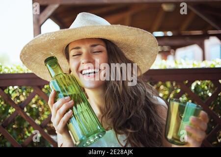 feliz ama de casa de moda en camisa blanca con botella de agua, vidrio y sombrero en la terraza. Foto de stock