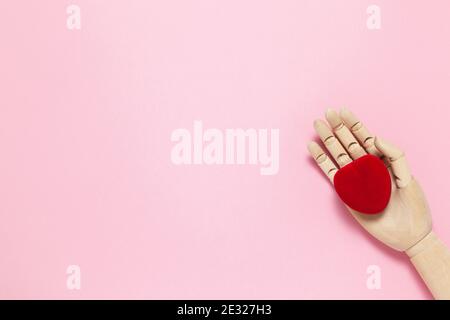 Mano de madera sostiene rojo pequeño terciopelo caja en forma de corazón para joyas sobre fondo rosa, espacio de copia, laico plano. 8 de marzo, 14 de febrero, cumpleaños, San Valentín, Foto de stock