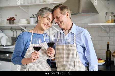 Feliz pareja de la vieja unión, bebiendo vino de pie en la cocina cocinando en casa.