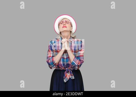 Oh mi Dios, por favor ayúdame. Retrato de esperanza mujer madura en moderno estilo casual con sombrero, anteojos de pie con los ojos cerrados y orando. Foto de stock