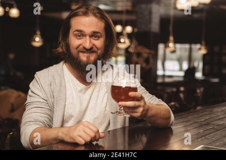 Alegre joven barbudo hombre sonriendo feliz a la cámara, sosteniendo una copa de deliciosa cerveza artesanal, espacio de copia. Hombre feliz disfrutando de una cerveza sabrosa en la loca Foto de stock