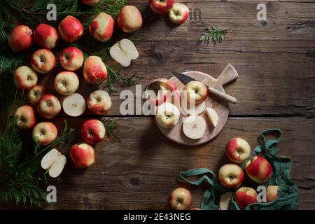 Vista superior de manzanas enteras frescas maduras y cortadas en rodajas rojas con tabla de cortar y cuchillo dispuesto en mesa de madera rústica con follaje verde