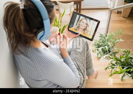 Mujer madura tomando café mientras que hace videollamada en digital tableta en casa