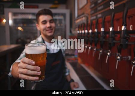 Barman feliz sosteniendo el vaso de cerveza a la cámara, trabajando en su bar Foto de stock