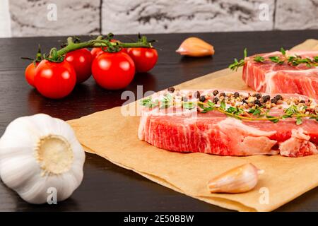 filetes de cerdo crudos recién picados con especias y tomillo en una hoja de pergamino, junto a tomates rojos maduros y ajo Foto de stock