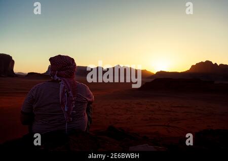 Un hombre árabe con keffiyeh jordano atado cabeza está sentado en la cima de una roca con vistas al desierto de Wadi Rum en Jordania. Está viendo la puesta de sol Foto de stock