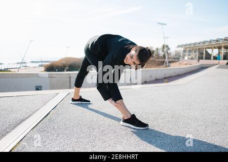 Longitud completa Determined joven Sportswoman en activo estirando piernas mientras que calienta para arriba en carretera en suburbio Foto de stock