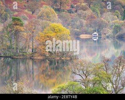El color otoñal se refleja en Rydal Water en una tranquila mañana de noviembre, con la icónica casa de barco visible en la orilla lejana. Foto de stock