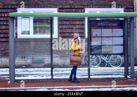 Mujer morena de mediana edad de pie en una parada de autobús, con una máscara protectora debido al virus de la corona, esperando a que su autobús la lleve a trabajar Foto de stock