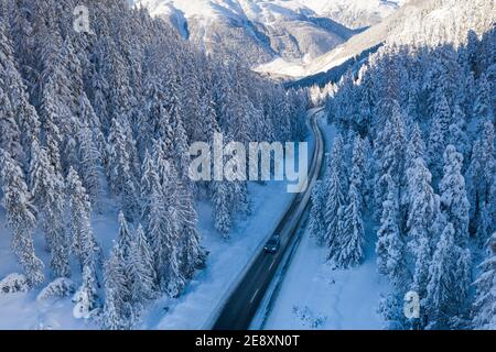 Coche viajando por carretera helada enmarcada por árboles cubiertos de nieve en el bosque de invierno, vista aérea, Suiza